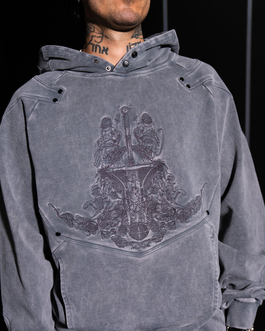 Person wearing a gray hoodie with a detailed design on the chest against a black background