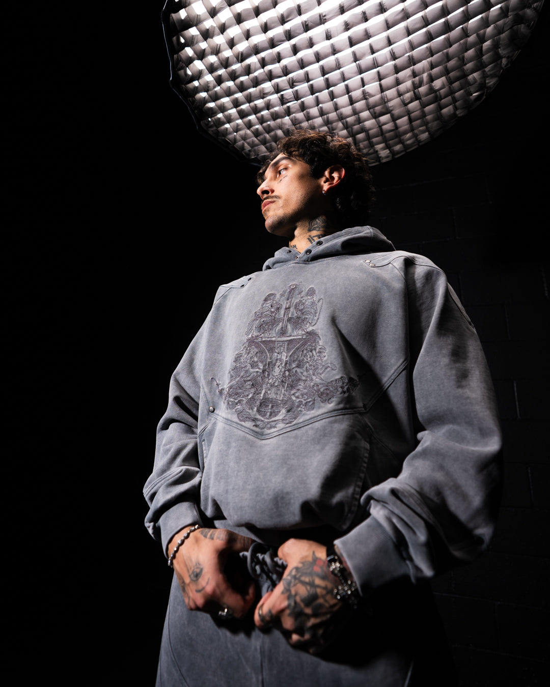 Person wearing a gray hoodie with a graphic design, standing against a black background with studio lighting.