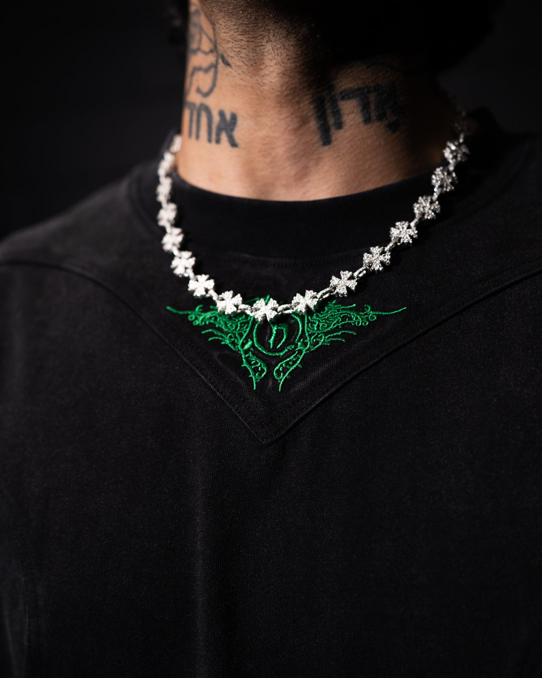 Person wearing a black shirt with green embroidery and a necklace on a dark background