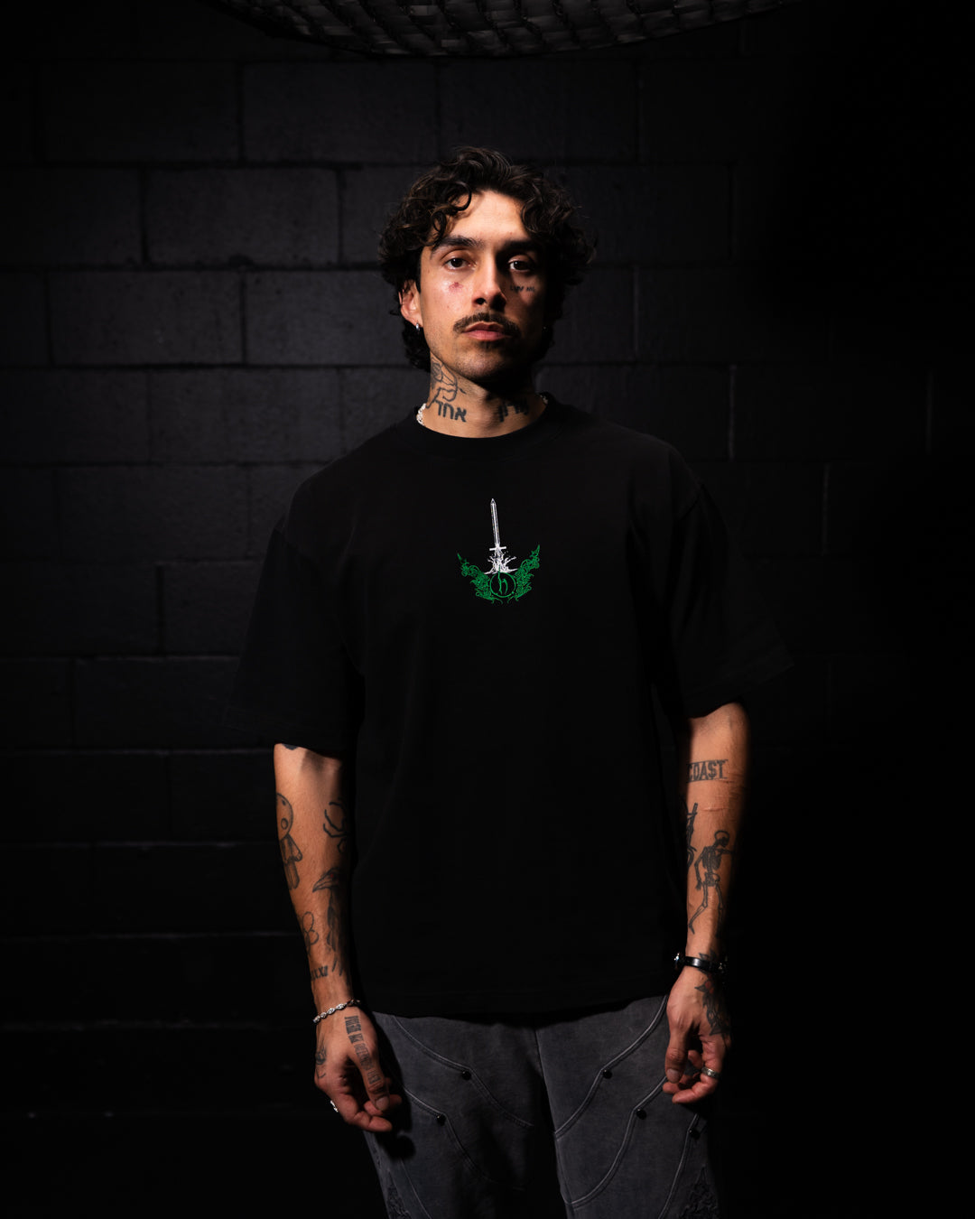 Person wearing a black t-shirt with a green graphic design against a dark brick wall.