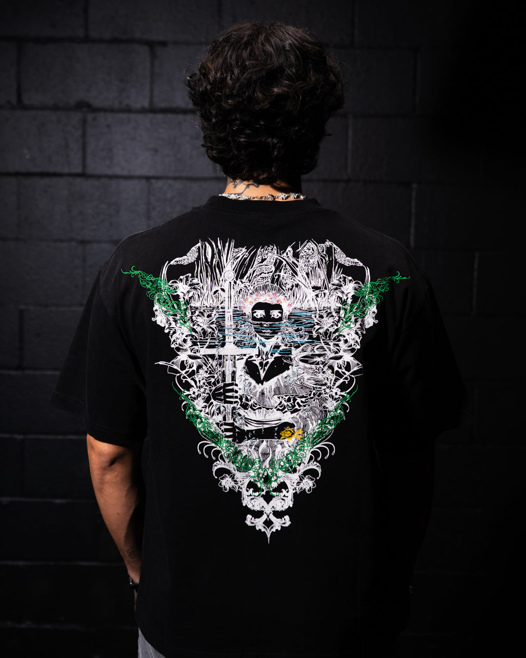 Person wearing a black t-shirt with a detailed design on the back against a dark brick wall.