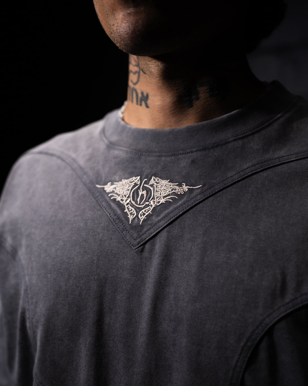 grey t-shirt with a decorative emblem on the collar against a dark background