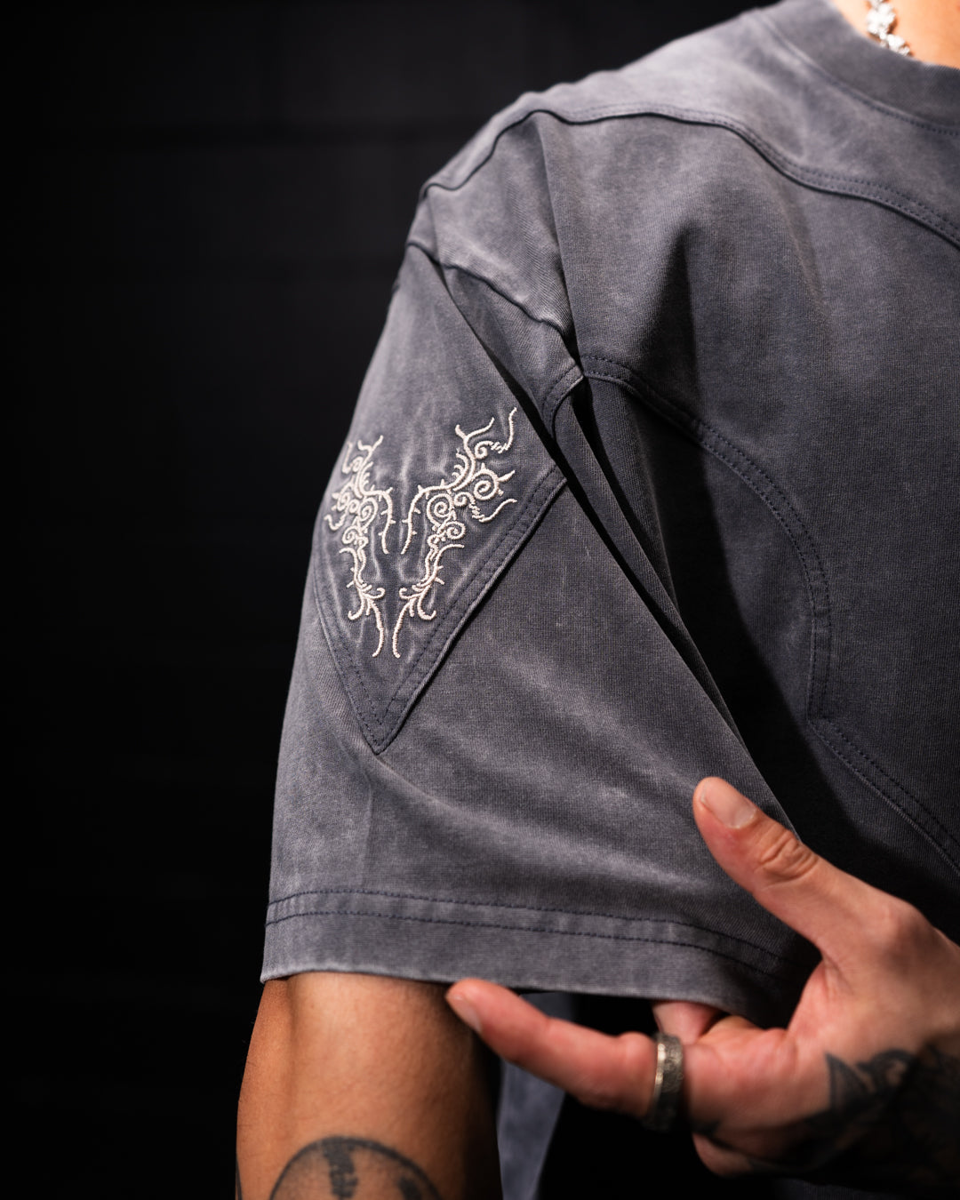 Person wearing a dark gray t-shirt with an embroidered design on the sleeve against a black background
