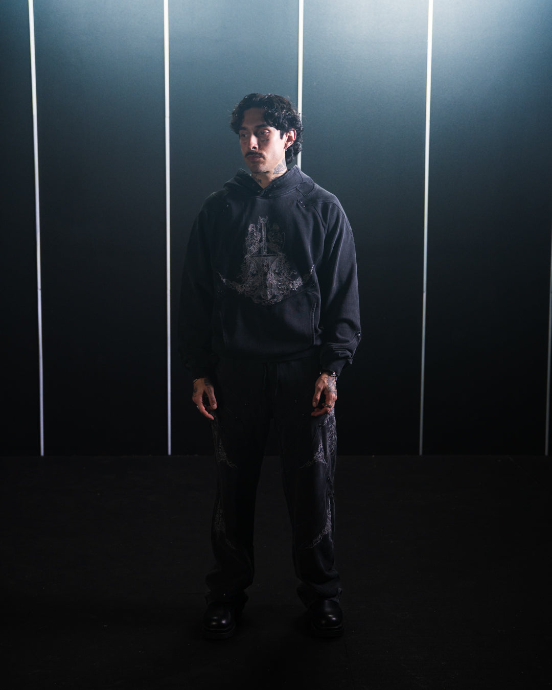 Person wearing a dark hoodie and pants standing against a dark background with vertical light strips.
