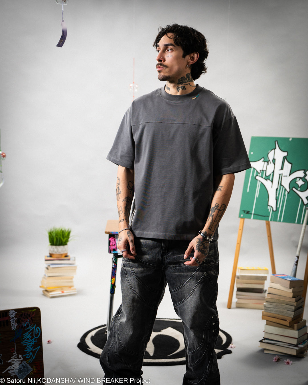 Man wearing a gray t-shirt and dark jeans standing in a room with books and a plant.