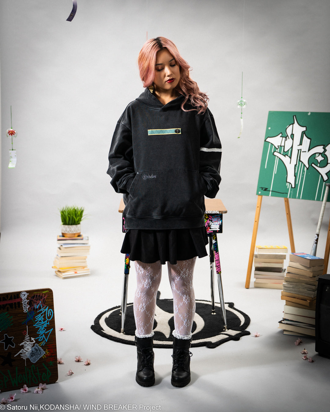 Person wearing a black hoodie and skirt in a room with books and a plant.