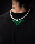 Person wearing a black shirt with green embroidery and a necklace on a dark background
