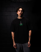 Person wearing a black t-shirt with a green graphic design against a dark brick wall.