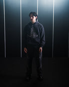 Person wearing a dark hoodie and pants standing against a dark background with vertical light strips.