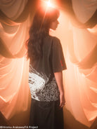 Person wearing a black  t-shirt with white graphics standing in front of draped fabric with warm lighting.