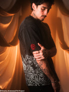 Man wearing a black shirt with white patterns and a red emblem, standing against a warm-toned curtain.