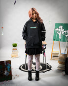 Person wearing a black hoodie and skirt in a room with books and a plant.