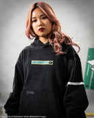 Person wearing a black hoodie with a logo patch on the front, standing against a gray background